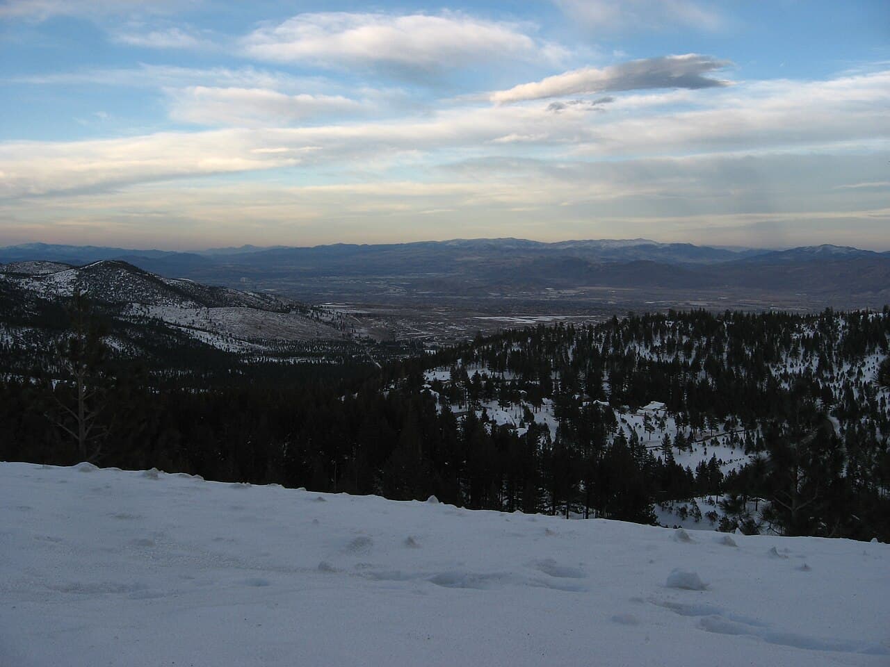 Snow-covered overlook on the Mount Rose Highway at dusk, looking down through pine forest toward the distant Truckee Meadows valley.